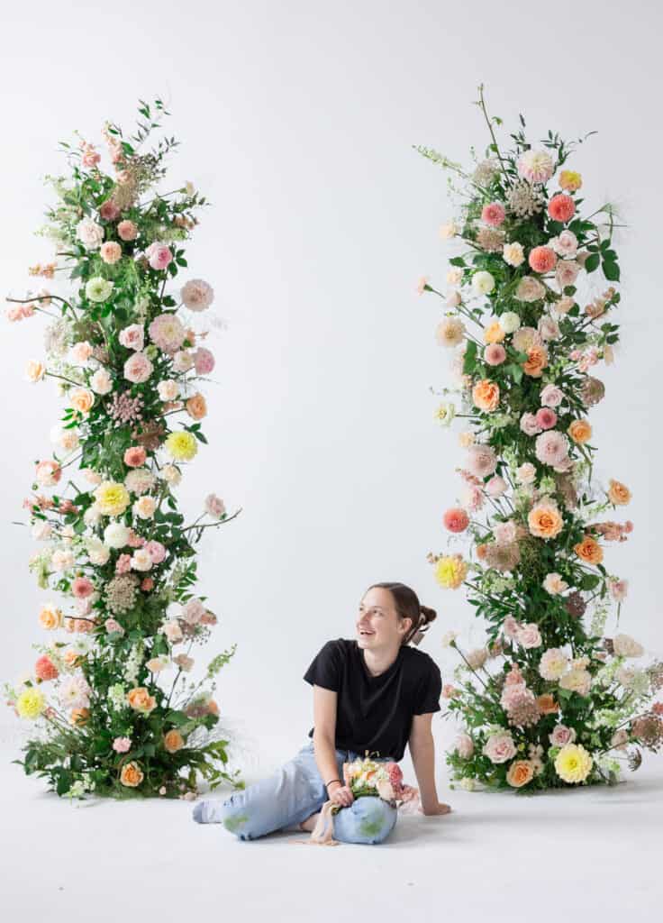 Caroline Hardy, a utah wedding florist, sits in front of an open arch, or two pillars of floral installments. The pillars have many colorful flowers on them. Caroline is holding a wedding bouquet with a pink ribbon. 