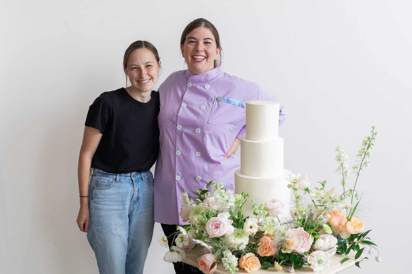 Caroline and Heather, a wedding florist and a wedding baker, pose in a hug. They are standing behind an antique white table that has a wedding cake and a flower cake meadow on it. They are Utah wedding vendors who like to collaborate and both own small businesses. 