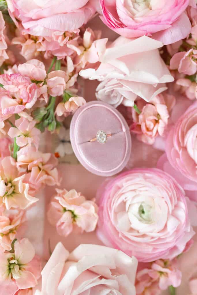 A diamond ring with a rose gold colored setting sits in an oval velvet case, surrounded by fresh pink flowers